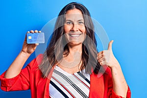 Young beautiful brunette businesswoman holding credit card over isolated blue background smiling happy and positive, thumb up