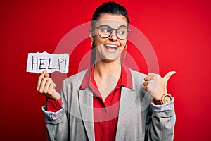 Young beautiful brunette businesswoman with problems holding paper with help message pointing and showing with thumb up to the