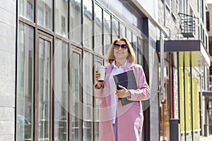 a young beautiful brunette businesswoman in sunglasses and a pink coat is walking along a city street