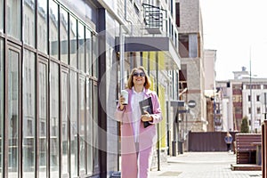 a young beautiful brunette businesswoman in sunglasses and a pink coat is walking along a city street