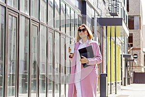 a young beautiful brunette businesswoman in sunglasses and a pink coat is walking along a city street