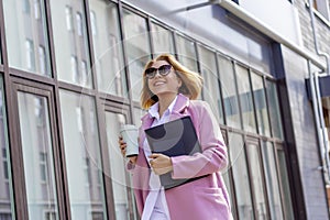 a young beautiful brunette businesswoman in sunglasses and a pink coat is walking along a city street