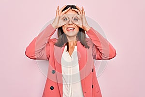 Young beautiful brunette businesswoman wearing elegant jacket over isolated pink background doing ok gesture like binoculars