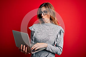 Young beautiful brunette businesswoman wearing glasses using laptop over red background scared in shock with a surprise face,