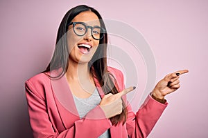 Young beautiful brunette businesswoman wearing jacket and glasses over pink background smiling and looking at the camera pointing