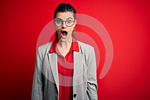 Young beautiful brunette businesswoman wearing jacket and glasses over red background afraid and shocked with surprise and amazed