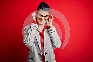 Young beautiful brunette businesswoman wearing jacket and glasses over red background covering ears with fingers with annoyed