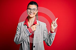 Young beautiful brunette businesswoman wearing jacket and glasses over red background smiling and looking at the camera pointing