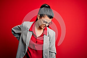 Young beautiful brunette businesswoman wearing jacket and glasses over red background Suffering of backache, touching back with