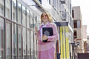 a young beautiful brunette businesswoman in a white blouse and light trousers and a pink coat is walking along a city street, in