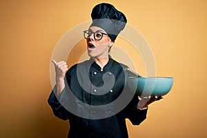 Young beautiful brunette chef woman wearing cooker uniform and hat holding bowl and whisk pointing and showing with thumb up to