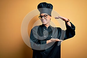 Young beautiful brunette chef woman wearing cooker uniform and hat over yellow background gesturing with hands showing big and