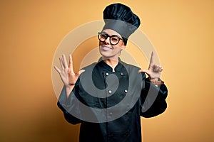 Young beautiful brunette chef woman wearing cooker uniform and hat over yellow background showing and pointing up with fingers