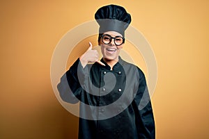 Young beautiful brunette chef woman wearing cooker uniform and hat over yellow background smiling doing phone gesture with hand