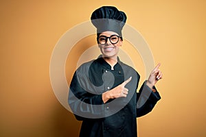 Young beautiful brunette chef woman wearing cooker uniform and hat over yellow background smiling and looking at the camera