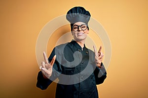 Young beautiful brunette chef woman wearing cooker uniform and hat over yellow background smiling looking to the camera showing