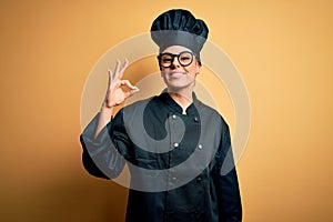 Young beautiful brunette chef woman wearing cooker uniform and hat over yellow background smiling positive doing ok sign with hand