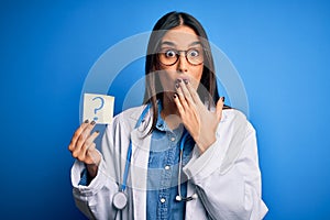 Young beautiful brunette doctor woman holding paper with question mark symbol message cover mouth with hand shocked with shame for