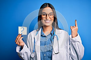 Young beautiful brunette doctor woman holding paper with question mark symbol message surprised with an idea or question pointing