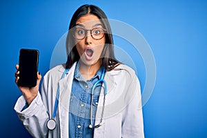 Young beautiful brunette doctor woman wearing glasses and coat holding smartphone scared in shock with a surprise face, afraid and