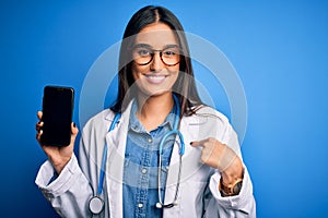 Young beautiful brunette doctor woman wearing glasses and coat holding smartphone with surprise face pointing finger to himself