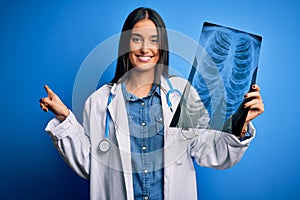 Young beautiful brunette doctor woman wearing stethoscope holding chest xray very happy pointing with hand and finger to the side