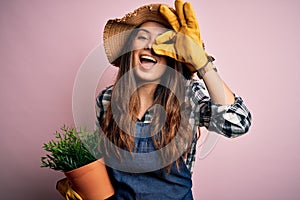 Young beautiful brunette farmer woman wearing apron and hat holding pot with plants with happy face smiling doing ok sign with