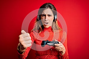 Young beautiful brunette gamer woman playing video game using joystic and headphones annoyed and frustrated shouting with anger,
