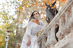 Beautiful young woman in a long white dress with brown horse outdoor.