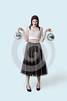 Young beautiful brunette girl in dark box pleated midi skirt posing in studio. Woman balancing holding two disco balls