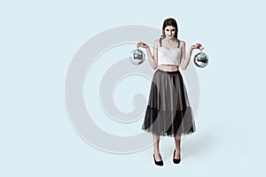 Young beautiful brunette girl in dark box pleated midi skirt posing in studio. Woman balancing holding two disco balls