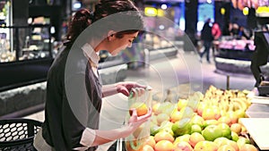 Young beautiful brunette girl in her 20`s picking out oranges into a plastic bag at the fruit and vegetable aisle in a