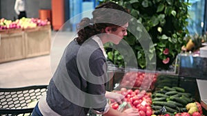 Young beautiful brunette girl in her 20`s picking out tomatoes into a plastic bag at the fruit and vegetable aisle in a