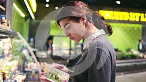 Young beautiful brunette girl in her 20`s trying to choose prepacked salad leaves in a grocery store.