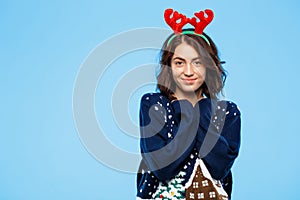 Young beautiful brunette girl in knited sweater and christmas reindeer antlers smiling over blue background.