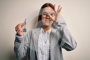 Young beautiful brunette house agent woman holding key home over white background with happy face smiling doing ok sign with hand