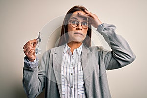 Young beautiful brunette house agent woman holding key home over white background stressed with hand on head, shocked with shame