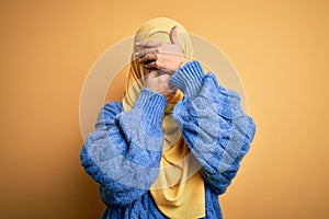 Young beautiful brunette muslim woman wearing arab hijab over isolated yellow background Covering eyes and mouth with hands,