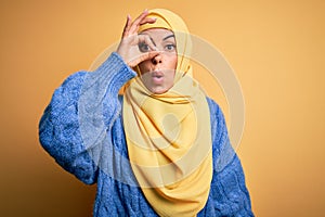 Young beautiful brunette muslim woman wearing arab hijab over isolated yellow background doing ok gesture shocked with surprised