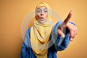 Young beautiful brunette muslim woman wearing arab hijab over isolated yellow background Pointing with finger surprised ahead,