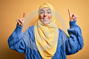 Young beautiful brunette muslim woman wearing arab hijab over isolated yellow background smiling amazed and surprised and pointing