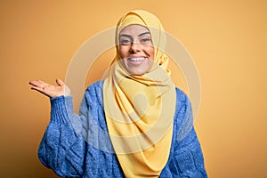 Young beautiful brunette muslim woman wearing arab hijab over isolated yellow background smiling cheerful presenting and pointing