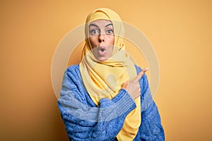 Young beautiful brunette muslim woman wearing arab hijab over isolated yellow background Surprised pointing with finger to the