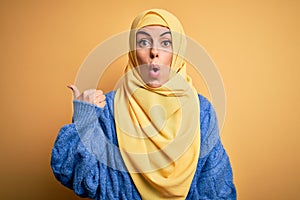 Young beautiful brunette muslim woman wearing arab hijab over isolated yellow background Surprised pointing with hand finger to