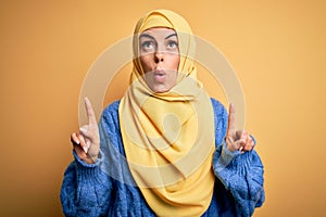 Young beautiful brunette muslim woman wearing arab hijab over  yellow background amazed and surprised looking up and