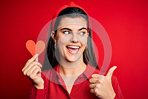 Young beautiful brunette romantic woman holding red heart paper for valentine day pointing and showing with thumb up to the side