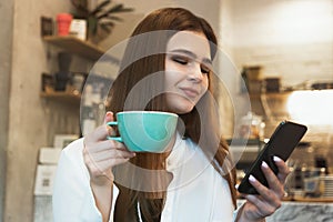Young beautiful brunette smiling woman drinks hot coffee in the cafe looks happy checking her smartphone