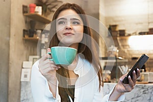 Young beautiful brunette smiling woman drinks hot coffee in the cafe looks happy holding smartphone in her hand