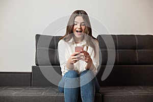 Young beautiful brunette smiling woman texting message in her smartphone sitting on the sofa in her appartment looks