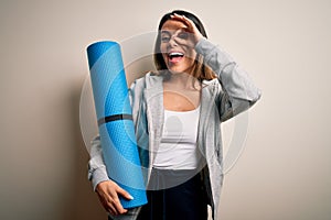 Young beautiful brunette sporty woman holding yoga mat over isolated white background with happy face smiling doing ok sign with